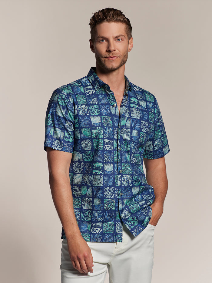 green leaf and ocean- grid print short-sleeve shirt and white pants, man with one hand in pocket, front view.