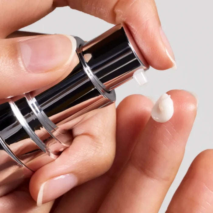 Hand dispensing white cream from shiny silver pump onto fingertip against plain background