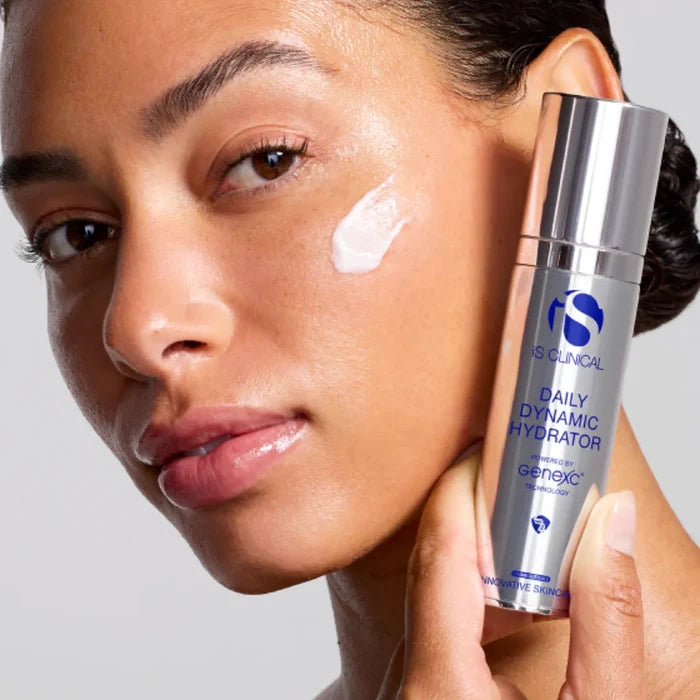 Close-up of woman with cream on cheek holding iS Clinical Daily Dynamic Hydrator silver bottle