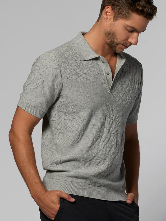 Man in Grey knit sweater polo, looking down with one hand in pocket, front view.