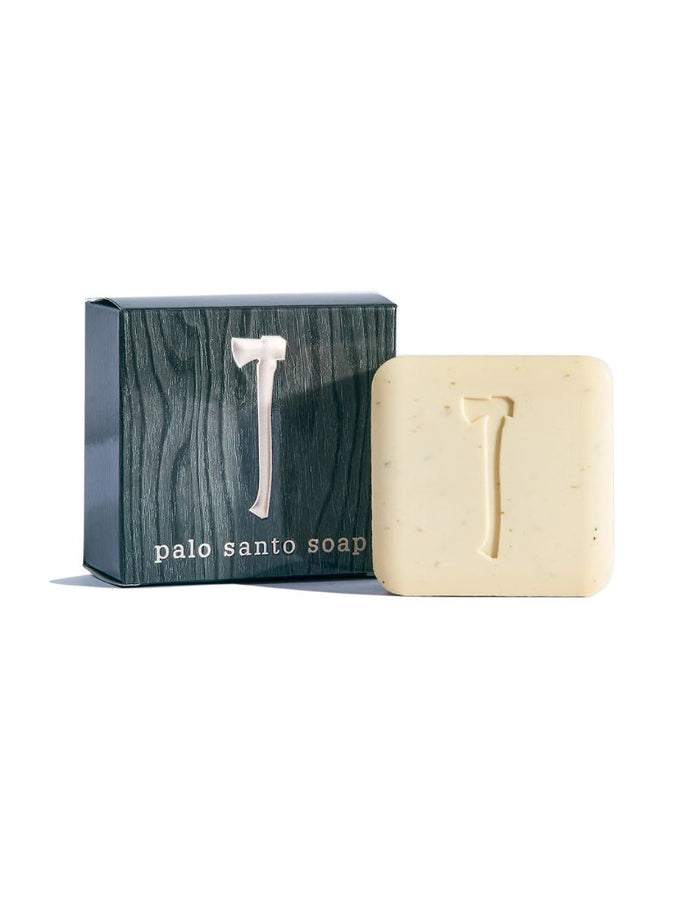 A bar of Palo Santo soap in a square cream-colored bar with a carved axe motif, displayed next to its dark green wood-grain textured packaging box with a silver axe logo.