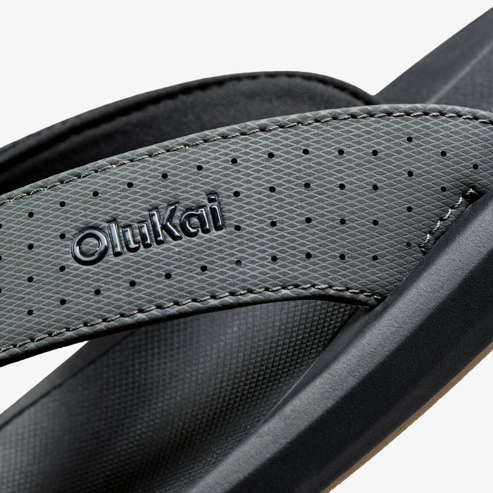 OluKai flip-flop with perforated gray textile strap and black sole, showing brand name and stitching details.