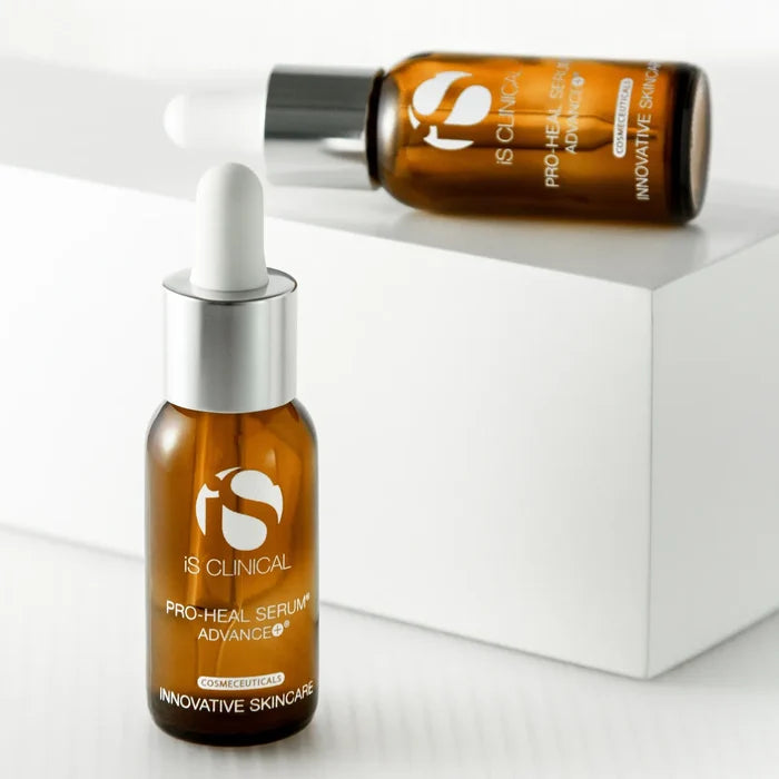Two amber glass bottles of iS Clinical Pro-Heal Serum Advance+ with white dropper caps on white geometric surfaces